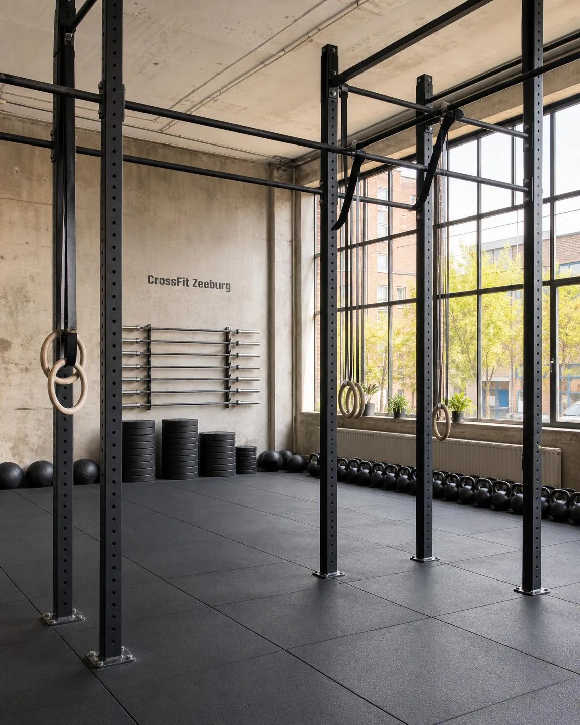 De trainingsvloer van CrossFit Zeeburg: betonnen wanden, zwarte rubbervloer, een stalen rig met ringen, barbells, bumperplates en kettlebells in daglicht.